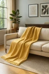 Koc bawełniany 150x200 cm w kolorze żółtym z frędzlami, leżący na beżowej sofie. Wyraźna struktura materiału, frędzle na dolnej krawędzi. W tle widoczna roślina doniczkowa oraz obrazki na ścianie, co tworzy przytulną atmosferę wnętrza.