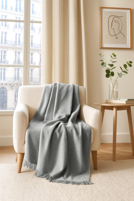 Koc bawełniany 150x200 w kolorze szarym, leżący na jasnym, tapicerowanym fotelu. Długi, z frędzlami na dolnej krawędzi, o gładkiej fakturze. W tle okno oraz jasne zasłony, co tworzy przytulną aranżację wnętrza.