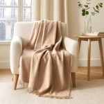 Koc bawełniany 200x220 w jasnym beżu, rozłożony na oparciu jasnoszarego fotela, z delikatnymi frędzlami na dolnej krawędzi. W tle widoczne okno oraz stół z rośliną w szklanym wazonie. Tło jasne i stonowane, podkreślające kolor koca.