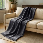 Koc bawełniany 150x200 cm w kolorze ciemnoszarym, z frędzlami. Produkt leży na jasnej kanapie, częściowo zwisając. Widoczny jest jego miękki materiał i tekstura. Koc w aranżacji wnętrza, w otoczeniu roślin oraz drewnianych mebli.