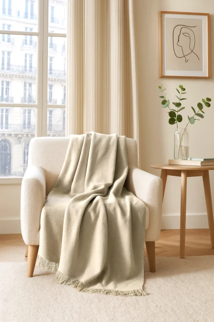Koc bawełniany 150x200 cm w kolorze kremowym leży na jasnym, miękkim fotelu. Produkt ma delikatne frędzle u dołu, a tło przedstawia jasne zasłony oraz stół z książkami i wazonem z rośliną. Całość w domowym, przytulnym wnętrzu.