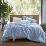 Pościel satynowa w odcieniu błękitnym 220x200, leżąca na łóżku z drewnianą ramą. Elemeny: narzuta, poduszki w różnych kształtach, dodatkowy koc. Otoczenie: naturalne światło zewnętrzne, roślinność w tle.