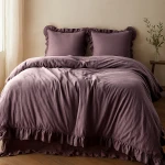 Pościel bawełniana 160x200 w kolorze fioletowym z falbanką, złożona na łóżku. Poduszki z falbanką w tym samym odcieniu. Tło jasne, drewniana podłoga, roślina w doniczce obok.