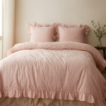 Pościel bawełniana 220x200 w kolorze różowym z falbanką, złożona na łóżku. W komplecie dwa poduszki z falbanką, o prostym, matowym wykończeniu. Tło w jasnych odcieniach, z delikatnymi detalami roślinnymi obok.