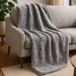 Puszysty koc dekoracyjny 150x200 cm w kolorze szarym z wielokolorowymi kropkami (zielonymi, białymi i fioletowymi) leży na szarej kanapie. Koc ma miękką, teksturowaną powierzchnię i jest częściowo zwinięty na oparciu.