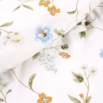 Pościel bawełniana 140x200 cm w białym kolorze z kolorowym kwiatowym wzorem. Widoczne różnokolorowe kwiaty, liście w odcieniach zieleni. Tkanina gładka, zbliżenie na detale wzoru.