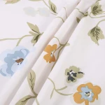 Pościel bawełniana 140x200 Meadowe w białym kolorze z kolorowym kwiatowym wzorem. Zbliżenie na tkaninę ukazującą detale niebieskich, pomarańczowych i zielonych kwiatów oraz liści, o gładkiej fakturze.