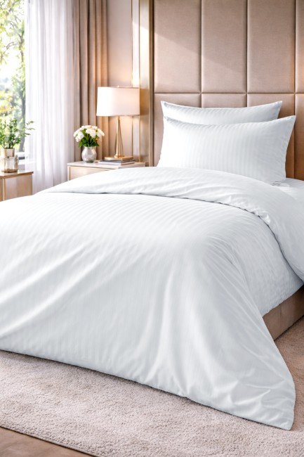 Pościel hotelowa bawełniana 140x200 w kolorze białym, z delikatnym wzorem w pasy o szerokości 1 cm. Złożona, leżąca na łóżku, z poduszką w komplecie. Tło w jasnym, stonowanym kolorze z elementami roślinnymi w aranżacji.