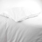 Pościel hotelowa bawełniana 220x200 w pasy 4-mm, biała, leżąca na podłożu. Widoczna poszewka na poduszkę z delikatnym przeszyciem. Faktura tkaniny gładka, z subtelnym połyskiem, na jednolitym tle. Rozmiar poduszki idealnie dopasowany do pościeli.