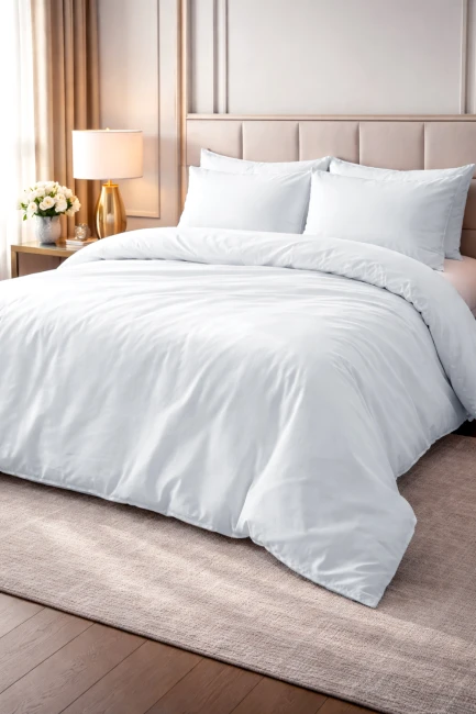 Pościel hotelowa bawełniana 220x200 cm, gładka, biała, leżąca na łóżku. W zestawie znajduje się poszewka na poduszkę oraz kołdra. Tło w jasnym wnętrzu z naturalnym drewnem i roślinnością, co podkreśla prostotę i elegancję produktu.