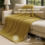 Koc żakardowy 200x220 w kolorze oliwkowym, z fakturą w formie drobnych, regularnych wzorów, leży na szarej, kanapie. W tle widoczna lampa na stoliku, co tworzy przytulny klimat aranżacji wnętrza.