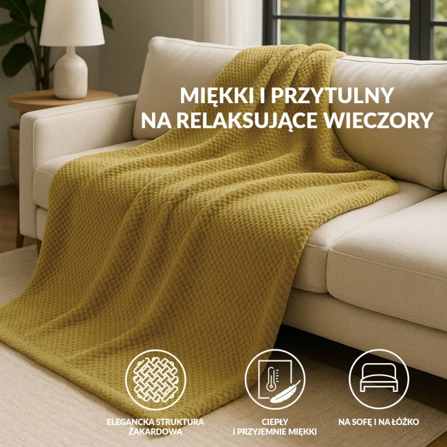 Koc żakardowy 200x220 w kolorze oliwkowym, z fakturą w formie drobnych, regularnych wzorów, leży na szarej, kanapie. W tle widoczna lampa na stoliku, co tworzy przytulny klimat aranżacji wnętrza.