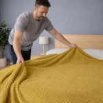 Koc żakardowy Amorro w kolorze oliwkowym o wymiarach 200x220 cm, z teksturowaną powierzchnią, rozłożony na łóżku. Mężczyzna trzyma koc, układając go na pościeli. Tło w stonowanej kolorystyce z drewnianym łóżkiem i lampką nocną.