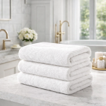 Ręcznik hotelowy 70x140 cm w kolorze białym, złożone na blacie, wykonany z miękkiego materiału o wysokiej chłonności. Zdjęcie przedstawia jasną łazienkę z naturalnym oświetleniem.