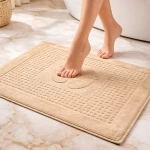 Dywanik łazienkowy 50x70 cm w kolorze cappuccino, z wypukłym wzorem w kształcie stóp. Ułożony na podłodze, z jedną stopą opartą na jego powierzchni. Zewnętrzna krawędź wykończona prostą lamówką, tekstura miękka i przyjemna w dotyku.