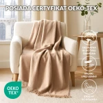 Koc bawełniany 200x220 w jasnym beżu, rozłożony na oparciu jasnoszarego fotela, z delikatnymi frędzlami na dolnej krawędzi. W tle widoczne okno oraz stół z rośliną w szklanym wazonie. Tło jasne i stonowane, podkreślające kolor koca.