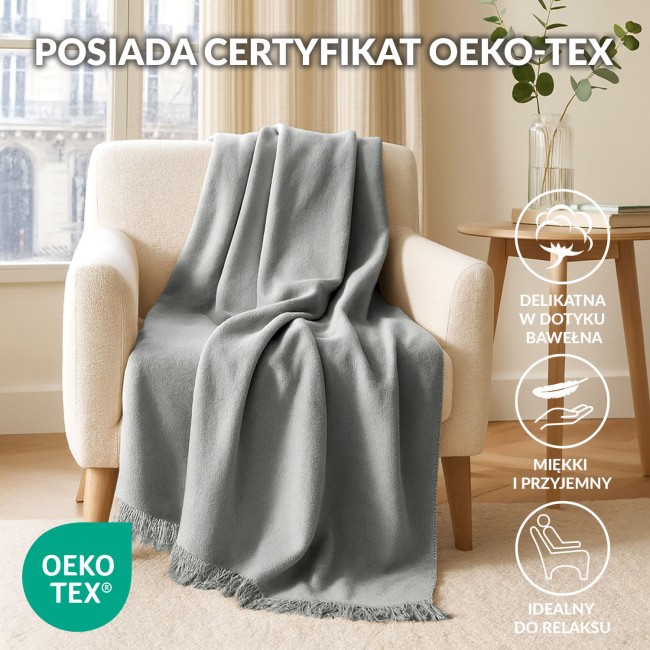 Koc bawełniany 150x200 cm w odcieniu szarości, z frędzlami na dolnej krawędzi. Produkt rozłożony na jasnej kanapie, nadaje przytulny charakter wnętrzu. W tle widoczne jasne zasłony i stół z dekoracyjną rośliną.