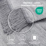 Koc bawełniany szary 150x200 cm, z frędzlami. Ułożony na płasko, z widoczną teksturą materiału. Frędzle wykończone estetycznie. Prostokątny kształt z gładkim wykończeniem brzegów. Dynie tło podkreśla szarość koca.