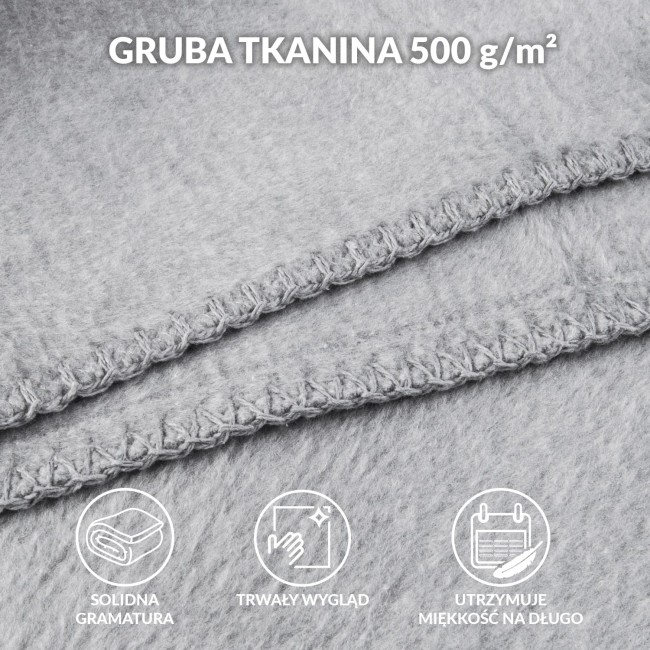 Koc bawełniany 150x200 w szarym kolorze, z detalami frędzli na krawędzi. Ułożony na powierzchni, widoczna tekstura materiału. Kąt ujęcia z boku ukazuje zagięcia oraz wykończenie, podkreślając jakość wykonania.