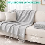 Koc bawełniany 150x200 cm w odcieniu szarości, z frędzlami na dolnej krawędzi. Produkt rozłożony na jasnej kanapie, nadaje przytulny charakter wnętrzu. W tle widoczne jasne zasłony i stół z dekoracyjną rośliną.