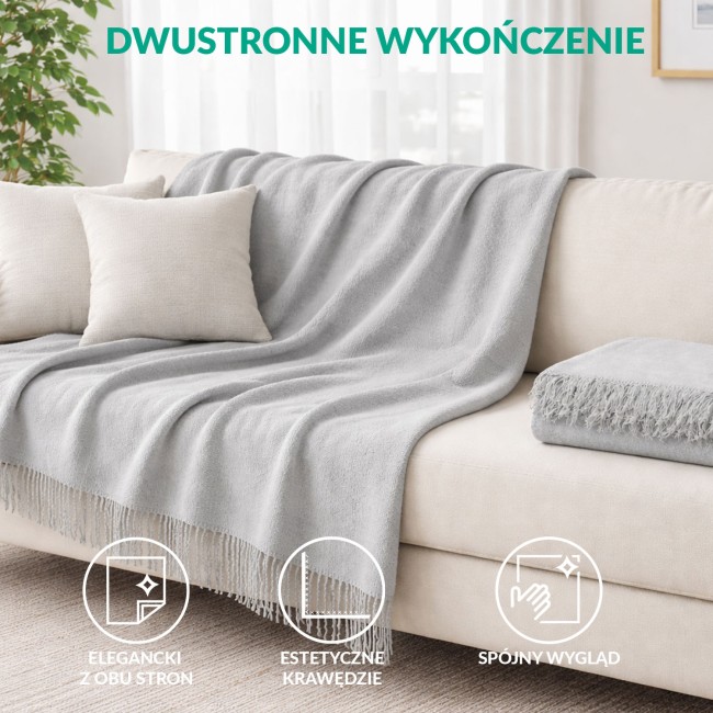 Koc bawełniany 200x220 cm w szarym kolorze, z frędzlami na jednym końcu, położony na jasnym fotelu. Tło stanowi jasne okno z roletami i delikatne, minimalistyczne elementy dekoracyjne. Koc sprawia wrażenie miękkiego i przytulnego.