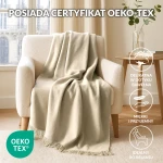 Koc bawełniany 150x200 cm w kolorze kremowym leży na jasnym, miękkim fotelu. Produkt ma delikatne frędzle u dołu, a tło przedstawia jasne zasłony oraz stół z książkami i wazonem z rośliną. Całość w domowym, przytulnym wnętrzu.