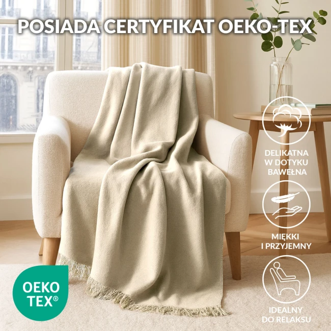 Koc bawełniany 150x200 cm w kolorze kremowym leży na jasnym, miękkim fotelu. Produkt ma delikatne frędzle u dołu, a tło przedstawia jasne zasłony oraz stół z książkami i wazonem z rośliną. Całość w domowym, przytulnym wnętrzu.