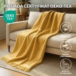 Koc bawełniany 150x200 cm w kolorze żółtym z frędzlami, leżący na beżowej sofie. Wyraźna struktura materiału, frędzle na dolnej krawędzi. W tle widoczna roślina doniczkowa oraz obrazki na ścianie, co tworzy przytulną atmosferę wnętrza.