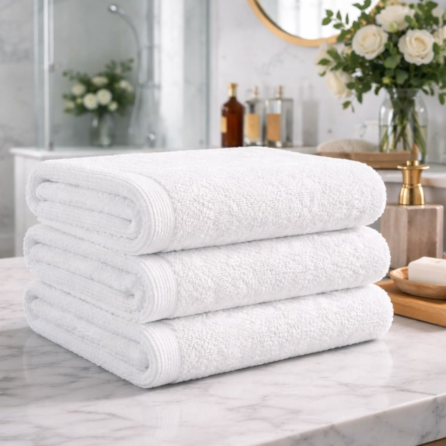 Biały ręcznik hotelowy 70x140 cm, gładki, o gramaturze 400 g/m², ułożony w stosie na marmurowym blacie, z widocznymi brzegami i teksturą materiału, w tle łazienka z roślinnością i dekoracjami.
