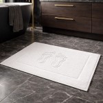 Biały dywanik łazienkowy 50x70 cm z wysokością runa 500 g/m², z wytłoczonym wzorem stóp. Ułożony na ciemnym kamiennym podłożu, w aranżacji łazienkowej, obok ciemnych mebli.