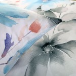Pościel satynowa 220x200 w kolorze białym z dużymi, pastelowymi niebieskimi kwiatami. Widoczne poduszki z delikatnym wzorem, w tle kwiaty w odcieniach szarości. Produkt leży na gładkiej powierzchni.
