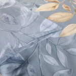 Pościel bawełniana 160x200 w kolorze niebieskim, ozdobiona beżowymi liśćmi. Poduszka z delikatnym wzorem liści, zbliżenie na fakturę materiału. Produkt leży na gładkim tle, ukazując detale nadruku.