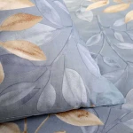 Pościel bawełniana w rozmiarze 160x200, w niebieskim kolorze z beżowym wzorem liści. Zbliżenie na poduszkę i narzutę, ukazujące delikatną teksturę materiału. Wzór liści jest subtelny, a całość ma matowe wykończenie.