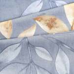 Pościel bawełniana 160x200 Sandleaf w odcieniu niebieskim z beżowymi liśćmi. Wzór liściowy na tkaninie o gładkiej fakturze, układ warstwowy materiału. Widoczne detale liści w beżowej i złotej kolorystyce.