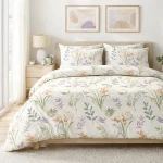 Pościel bawełniana 140x200 w kremowym kolorze z botanicznym wzorem kwiatowym, układająca się na łóżku z poduszkami. Widoczne delikatne detale roślinne w odcieniach zieleni, fioletu i pomarańczy, na tle jasnym. Ułożona na drewnianym łóżku w jasnym wnętrzu.