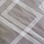 Pościel bawełniana 160x200 w odcieniu szarym z minimalistycznym, geometrycznym wzorem białych linii, widoczna na tle. Produkt leży płasko, ukazując detale faktury materiału oraz wyraźne linie tworzące prostokąty i krzyżujące się.