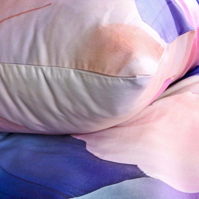Pościel satynowa 140x200 cm Aurora w białe i niebieskie pastelowe kwiaty. Widoczna poduszka z delikatnym wzorem w odcieniach różu i fioletu. Materiał gładki z połyskiem, ułożony na łóżku.