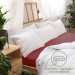 Prześcieradło jersey z gumką w kolorze bordowym, rozmiar 70x160 cm, rozłożone na łóżku z białymi poduszkami i narzutą. Tło to jasne zasłony oraz roślinność w doniczce, co tworzy przytulny klimat.