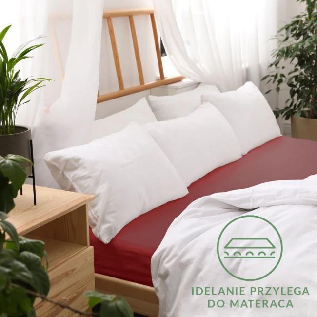 Prześcieradło jersey z gumką w kolorze bordowym, rozmiar 70x160 cm, rozłożone na łóżku z białymi poduszkami i narzutą. Tło to jasne zasłony oraz roślinność w doniczce, co tworzy przytulny klimat.