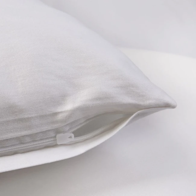 Poszewka satynowa 80x80 w kolorze białym, leżąca na jednolitym tle. Wykonana z gładkiego materiału z zamkiem błyskawicznym, co pozwala na łatwe zakładanie i zdejmowanie. Szczegół widać w dolnym rogu poszewki.