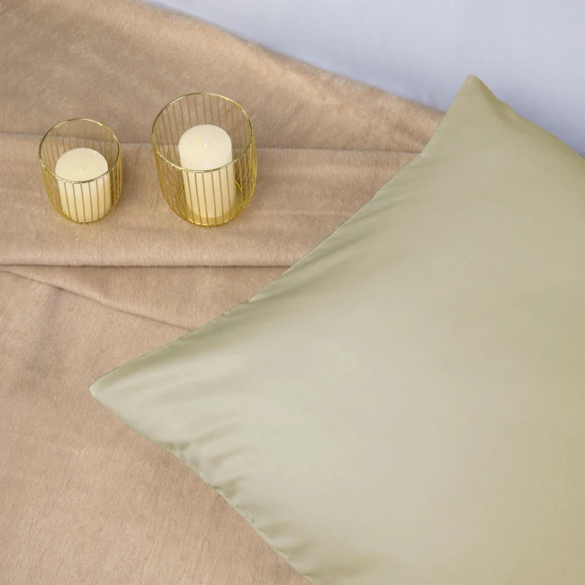 Poszewka satynowa 80x80 w kolorze kremowym, leżąca na jasnym tle. Widoczna gładka faktura materiału, bez wzorów. Obok stoją dwa świeczniki z żółtymi świecami. Tło proste, minimalne, skupione na produkcie.