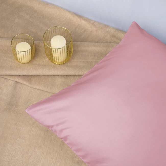 Poszewka satynowa 80x80 w kolorze wrzosowym, leżąca na jasnym, miękkim tle. Widoczne dwa świeczniki z przezroczystymi świecami. Zbliżenie na gładką fakturę poszewki, bez widocznych napisów lub zdobień.