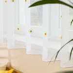 Firana BELLIS z etaminu, haftowana w stokrotki, biała, z ołowianką. Zawieszona w oknie, prezentuje delikatny wzór z żółtymi środkami kwiatów, z krawędzią wykończoną na gładko. Wymiary 300 x 160 cm.