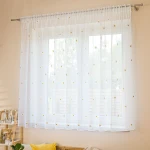 Firana BELLIS o wymiarach 300 x 160 cm, wykonana z etaminy, haftowana w stokrotki, mocowana na taśmie. Biała, z ołowianką, wisząca na karniszu, delikatnie przepuszczająca światło. Tło jasne, minimalistyczne, z elementami dekoracyjnymi w aranżacji.