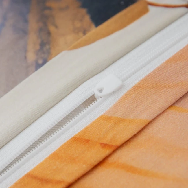 Zbliżenie na zamek błyskawiczny poszewki pościeli, z białym suwakiem i osłoną w kolorze kremowym. Widoczna tekstura materiału oraz delikatne zabarwienie w odcieniach beżu i pomarańczy. Poszewka leży na gładkiej powierzchni.