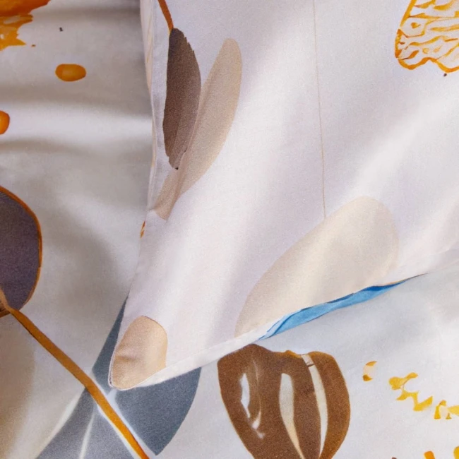 Pościel satynowa 200x200 cm w kolorze kremowym z niebieskimi motylami. Zbliżenie na poduszkę z delikatnym wzorem w odcieniach brązu i błękitu. Materiał gładki, błyszczący. Produkt leżący na płaskiej powierzchni. W tle neutralne, jasne kolory.