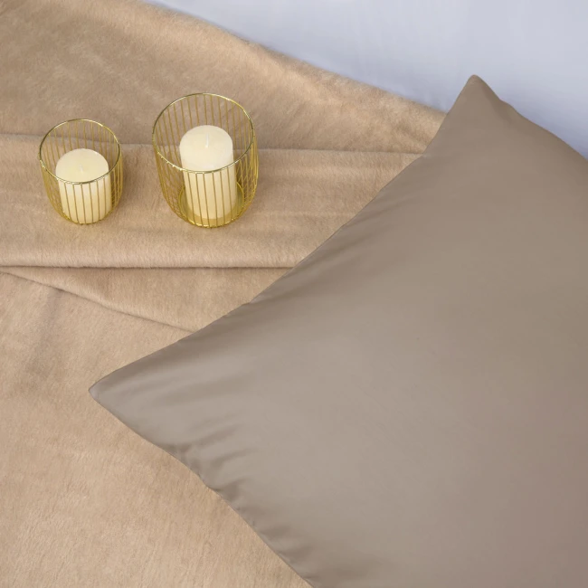 Poszewka satynowa w kolorze ciemnego beżu o wymiarach 70x80 leży na jasnym, miękkim tle. Produkt ma gładką, błyszczącą powierzchnię. Obok znajdują się dwa szklane świeczniki w złotej metalowej oprawie z jasnymi świecami wewnątrz.