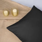 Poszewka satynowa 70x80 w kolorze czarnym leży na miękkim, beżowym tle. Pokazuje gładką powierzchnię, podkreślając elegancki wygląd. Obok znajdują się dwa świeczniki z białymi świecami, tworzące przytulną aranżację.