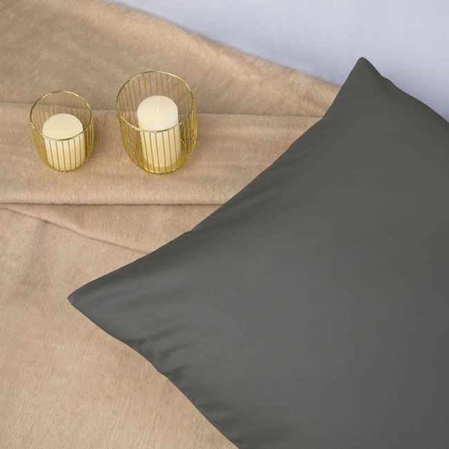 Poszewka satynowa 70x80 w kolorze szarym, leżąca na beżowym tle. Produkt ma gładką, błyszczącą fakturę. W tle widoczne są dwa szklane świeczniki w złotych osłonkach z jasnymi świecami.