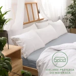 Prześcieradło jersey z gumką 60x120 w kolorze popiel na łóżku, z lewej strony widoczne białe poduszki. Produkt leży na materacu, z delikatnym grubym materiałem, który idealnie przylega. W tle rośliny i drewniane elementy, logo i tekst w dolnym rogu.